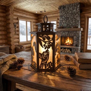 May include: A decorative brown metal lantern with a bird and floral cut-out design. The lantern is illuminated from within, casting a warm glow. It sits on a wooden table with pine cones and a blanket.