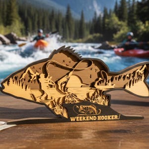 May include: A layered wooden fish-shaped art piece with a fishing scene, mountains, and the text "WEEKEND HOOKER". The art is in front of a river with kayakers and trees.