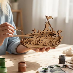 May include: A wooden tank model with intricate cut-out details, featuring soldiers and a flag. The model is being painted with a brush, showcasing the natural wood color. Paint pots and a palette are visible on the wooden table.