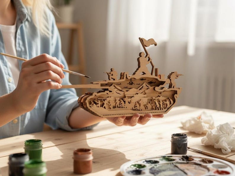 May include: A wooden tank model with intricate cut-out details, featuring soldiers and a flag. The model is being painted with a brush, showcasing the natural wood color. Paint pots and a palette are visible on the wooden table.