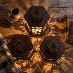 May include: Three dark wooden lanterns with a hexagonal design, each featuring a deer silhouette scene illuminated from within. The lanterns are arranged on a wooden surface with a cup of coffee, pinecones, and a blanket.
