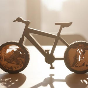 May include: A wooden bicycle sculpture with intricate cut-out designs in the wheels, depicting a mountain biking scene. The bicycle is brown and has a detailed frame, handlebars, seat, and pedals. The wheels feature a detailed forest scene.