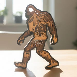 May include: A wooden Bigfoot figure, intricately layered with a detailed landscape design visible through the cutout silhouette. The figure is walking, with a light brown color and dark brown edges, and is placed on a wooden surface.
