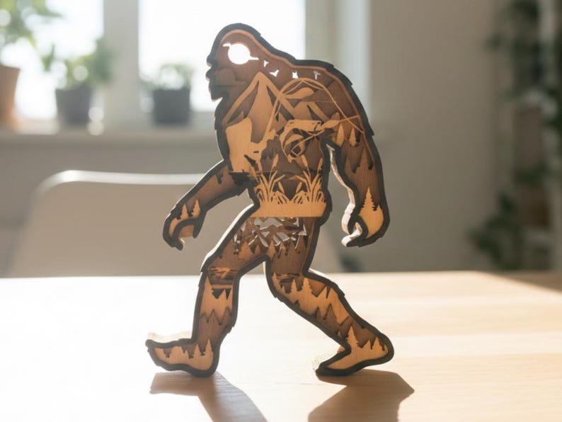 May include: A wooden Bigfoot figure, intricately layered with a detailed landscape design visible through the cutout silhouette. The figure is walking, with a light brown color and dark brown edges, and is placed on a wooden surface.