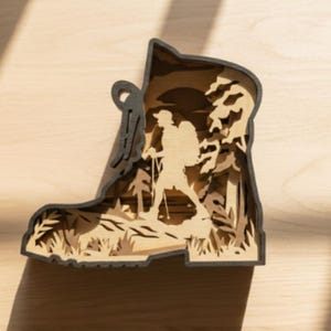 May include: A layered wooden art piece in the shape of a hiking boot. The interior depicts a hiker with a backpack and trekking poles in a forest scene. The boot's outline is dark gray, and the interior layers are light brown.