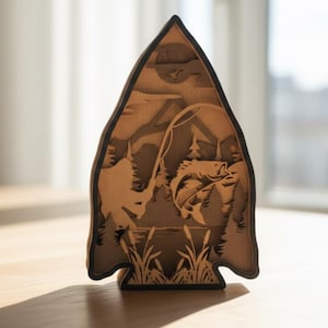 May include: A wooden layered art piece in the shape of an arrowhead, depicting a fishing scene. The artwork features a fish jumping out of the water, a fisherman, trees, and a lake. The piece is made of light brown wood with a dark brown border.