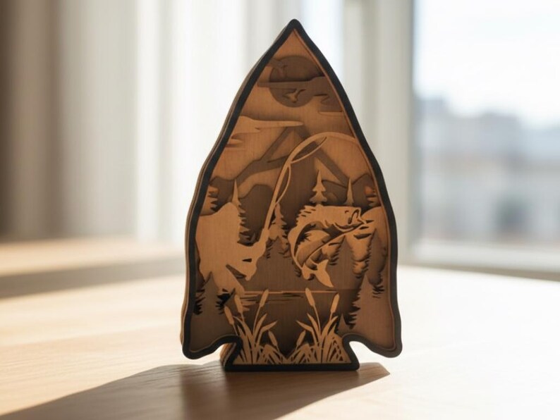 May include: A wooden layered art piece in the shape of an arrowhead, depicting a fishing scene. The artwork features a fish jumping out of the water, a fisherman, trees, and a lake. The piece is made of light brown wood with a dark brown border.