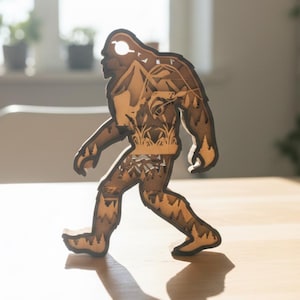 May include: A wooden Bigfoot figure, intricately cut with mountain and forest details. The brown figure is walking, with a hole at the top for hanging. The design is layered, creating a 3D effect. The figure is on a light-colored surface.
