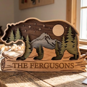 May include: A wooden bear-shaped sign with a mountain and forest scene, a moon, and stars. The bear is walking on a log that reads "THE FERGUSONS". The sign is made of layered wood with natural wood tones and green accents.