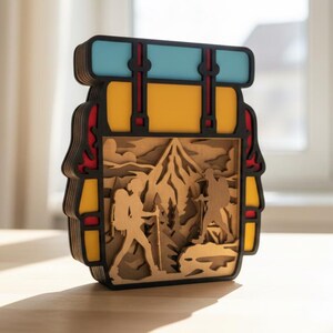 May include: A layered wooden art piece depicting a backpack with a mountain scene. The backpack has a yellow body, black trim, red accents, and a blue top. The interior shows hikers and trees.