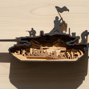 May include: A wooden layered art piece depicting a tank with soldiers and a flag. The tank is black with a light brown interior, featuring silhouettes of soldiers and a waving flag. The artwork is set against a light wood background.