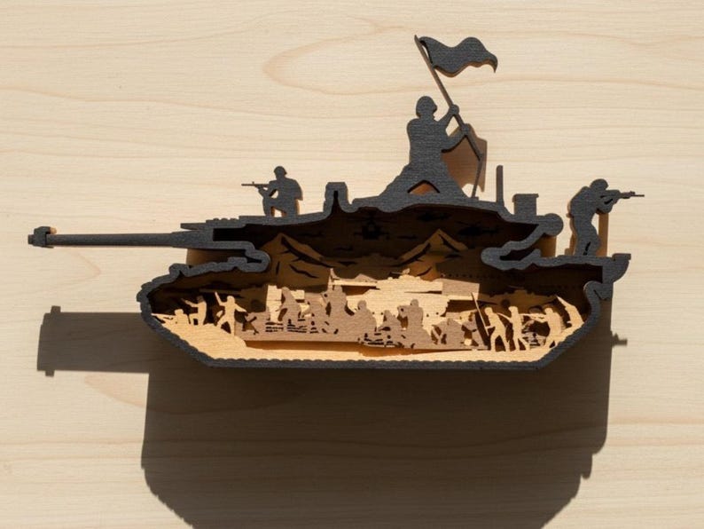May include: A wooden layered art piece depicting a tank with soldiers and a flag. The tank is black with a light brown interior, featuring silhouettes of soldiers and a waving flag. The artwork is set against a light wood background.