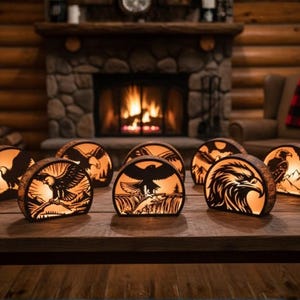 Op de afbeelding: Een verzameling verlichte houten lampen met adelaarsilhouetten. Elke lamp toont een ander adelaarsontwerp en straalt een warm, oranje licht uit. De lampen staan op een houten tafel, met een open haard op de achtergrond.
