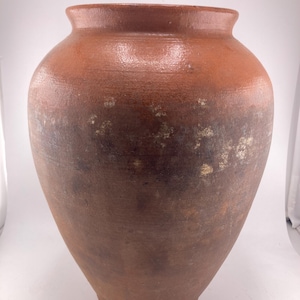 Puede incluir: Una jarra grande de cer&aacute;mica de estilo antiguo, con un cuerpo ancho y redondeado y un cuello estrecho. La jarra es de color marr&oacute;n rojizo con sombras m&aacute;s oscuras hacia la parte inferior y algunas manchas m&aacute;s claras. La superficie parece sin esmaltar.