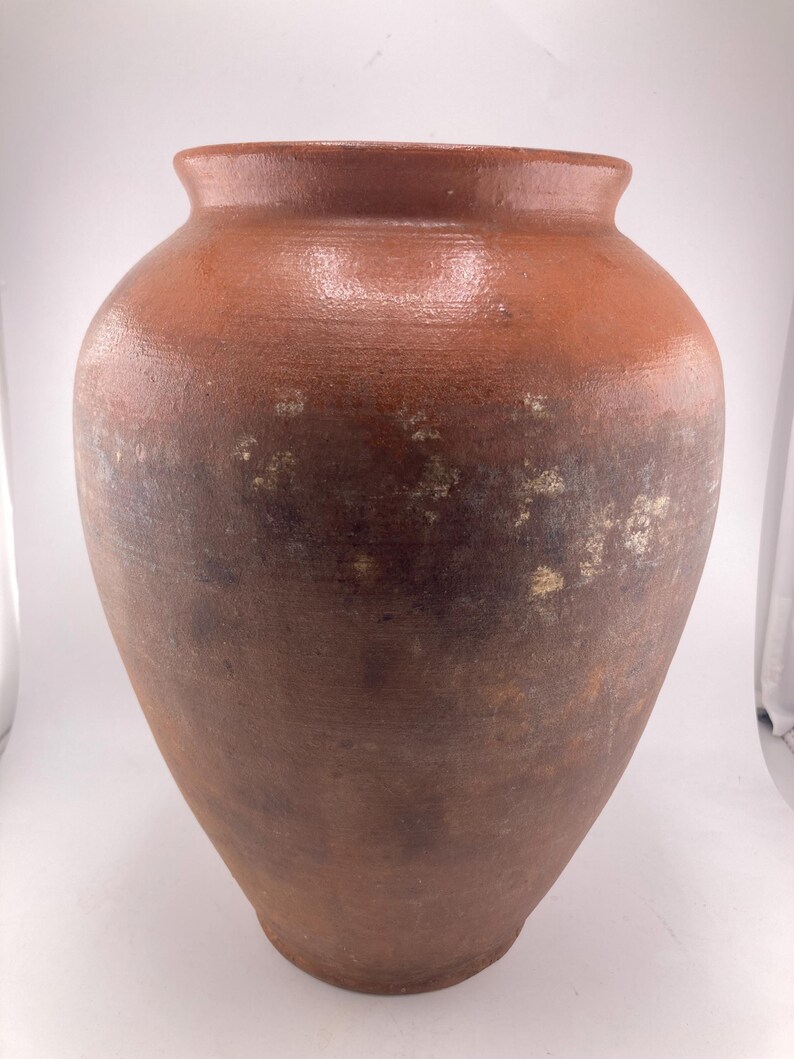 Puede incluir: Una jarra grande de cer&aacute;mica de estilo antiguo, con un cuerpo ancho y redondeado y un cuello estrecho. La jarra es de color marr&oacute;n rojizo con sombras m&aacute;s oscuras hacia la parte inferior y algunas manchas m&aacute;s claras. La superficie parece sin esmaltar.