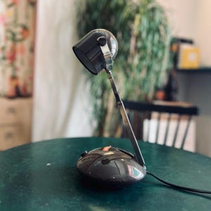 Könnte beinhalten: Eine dunkelgraue Schreibtischlampe mit einem abgerundeten Sockel und einem zylindrischen Kopf. Die Lampe verfügt über einen verstellbaren Metallarm, der den Sockel mit dem Leuchtengehäuse verbindet. Ein schwarzes Kabel führt vom Sockel weg.