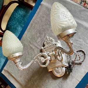 Może przedstawiać: Zabytkowy kinkiet ścienny z dwoma kloszami z matowego szkła. Kinkiet ma srebrną metalową ramę z ozdobnymi detalami. Klosze mają kształt stożka z kwiatowym wzorem. Kinkiet posiada przewód zasilający i wtyczkę.