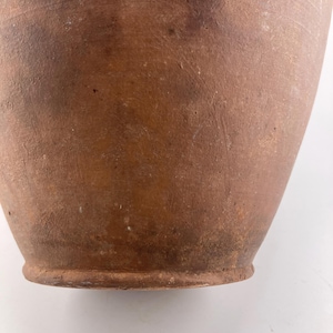 Puede incluir: Primer plano de un jarr&oacute;n de estilo antiguo, color terracota. El jarr&oacute;n tiene una base redondeada y un cuerpo ligeramente ensanchado. La superficie muestra variaciones de color y textura, con manchas m&aacute;s oscuras e imperfecciones visibles.