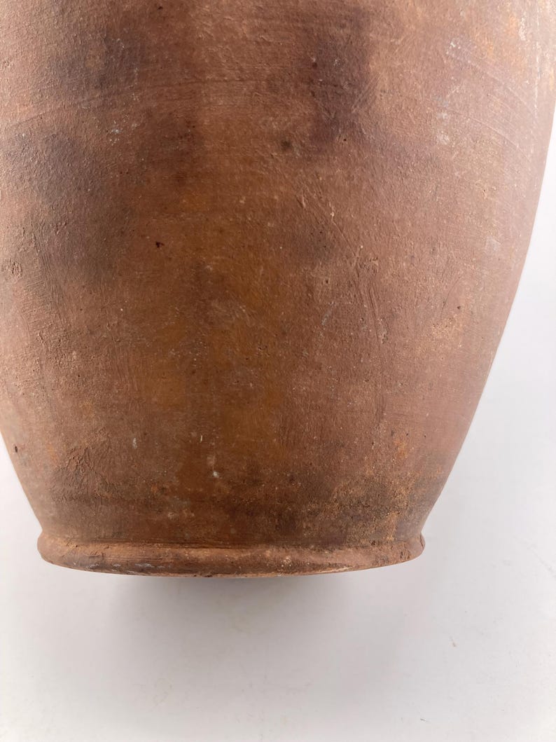 Puede incluir: Primer plano de un jarr&oacute;n de estilo antiguo, color terracota. El jarr&oacute;n tiene una base redondeada y un cuerpo ligeramente ensanchado. La superficie muestra variaciones de color y textura, con manchas m&aacute;s oscuras e imperfecciones visibles.