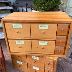 May include: A light brown wooden cabinet with multiple drawers and small brass knobs. The cabinet has labels on the drawers with text in a foreign language. The cabinet is stacked on top of another similar cabinet.