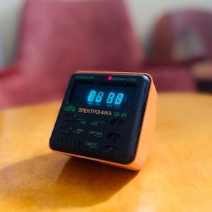 May include: A small, orange and black digital alarm clock with a blue LED display showing "88:88". The clock has buttons and text in a foreign language, including "Alfa Elektronika 12-41". It sits on a wooden surface.