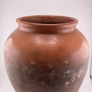 Puede incluir: Una jarra grande de cer&aacute;mica de estilo antiguo con una abertura ancha y un cuerpo redondeado. La jarra es de un color marr&oacute;n tierra c&aacute;lido con sombras m&aacute;s oscuras cerca de la base y manchas m&aacute;s claras. La superficie tiene una textura ligeramente rugosa.