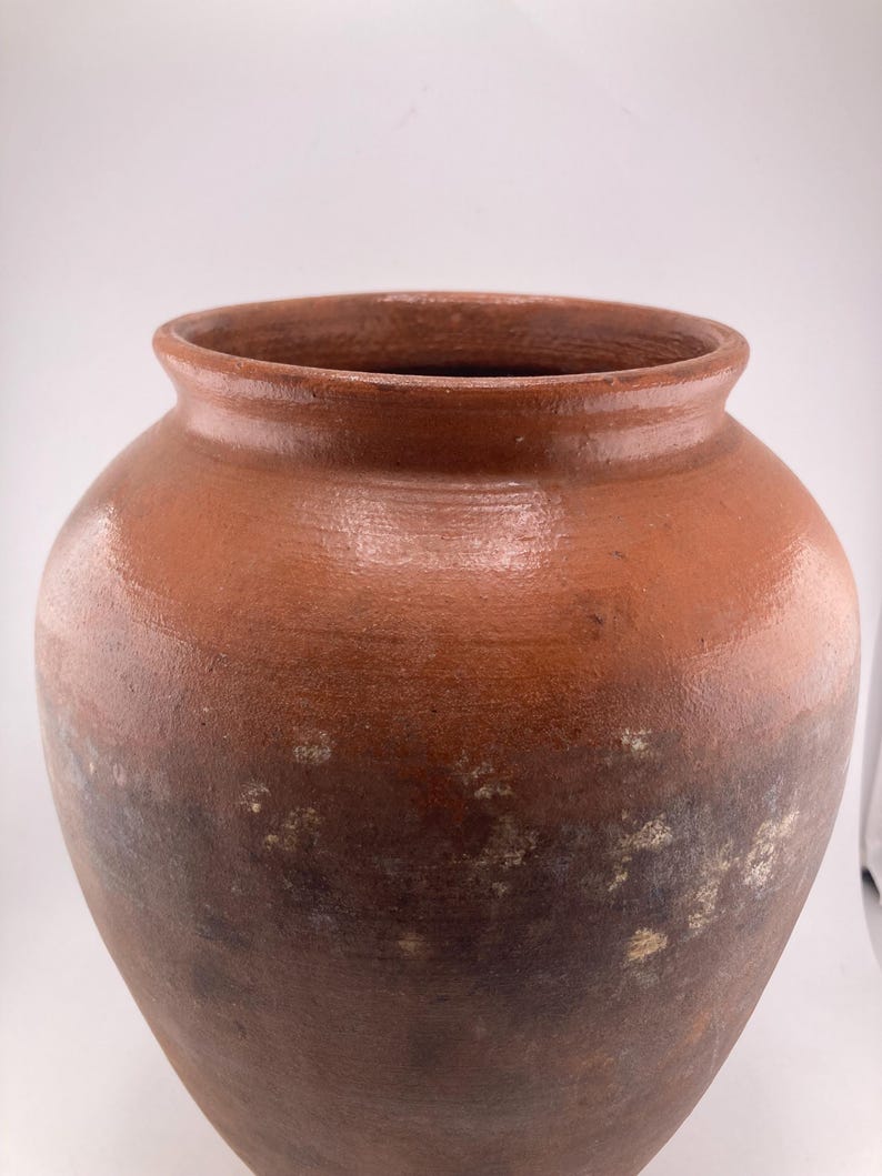 Puede incluir: Una jarra grande de cer&aacute;mica de estilo antiguo con una abertura ancha y un cuerpo redondeado. La jarra es de un color marr&oacute;n tierra c&aacute;lido con sombras m&aacute;s oscuras cerca de la base y manchas m&aacute;s claras. La superficie tiene una textura ligeramente rugosa.