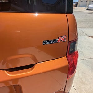 May include: Rear view of an orange vehicle with a black rear bumper. The vehicle has a black rear window and a red and black "TOAST R" emblem.