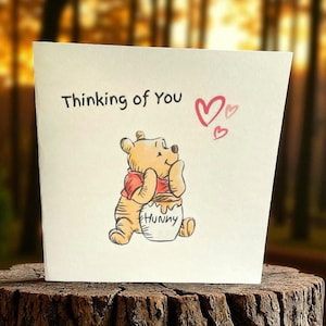 May include: A greeting card with the text "Thinking of You" and a drawing of Winnie the Pooh hugging a honey pot. The card features two pink hearts and is set against a blurred background.