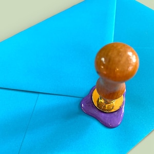 May include: A wooden handled wax seal stamp with a gold base, pressing into a purple wax seal on a bright blue envelope. The words "Seal & Send" are in the upper left corner.