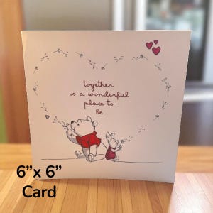 Op de afbeelding: Een kaart van 15 cm x 15 cm met Winnie de Poeh en Knorretje. De illustratie toont de personages die paardenbloemzaden blazen, omringd door een hartvormig ontwerp. De kaart bevat de tekst "together is a wonderful place to be".
