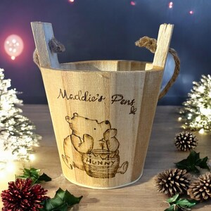 Könnte beinhalten: Hellbrauner Holzeimer mit Winnie-the-Pooh-Design und dem Text "Maddie's Pens". Der Eimer hat Holzgriffe mit Seildetails. Die Illustration zeigt Winnie Puuh und Ferkel mit einem Honigtopf.