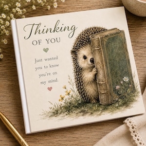 Può includere: Un biglietto d'auguri quadrato con l'illustrazione di un riccio che sbuca da dietro un libro antico verde. Il testo recita: Thinking OF YOU, Just wanted you to know you're on my mind. Sono presenti piccoli cuori verdi e rosa e fiori gialli.
