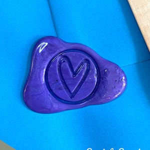 May include: A purple wax seal with a heart design inside a circle, on a blue background. The text "Seal & Send" is written in white script below the seal. The seal has a glossy finish.