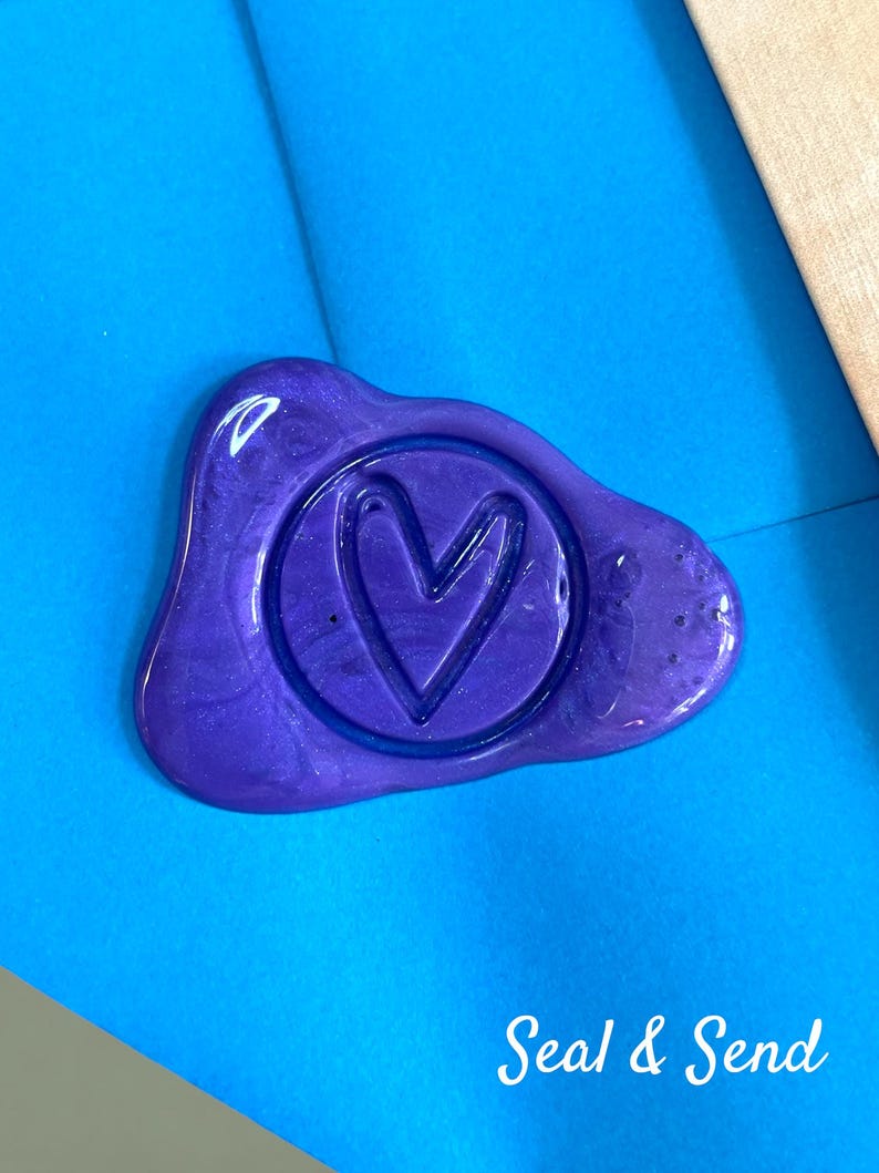 May include: A purple wax seal with a heart design inside a circle, on a blue background. The text "Seal & Send" is written in white script below the seal. The seal has a glossy finish.