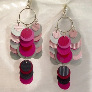 May include: A pair of dangle earrings with multiple layers of pink, silver, and black circles. The earrings are made of felt and have a silver hook closure.