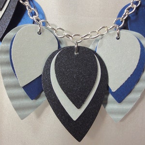 May include: A silver chain necklace with layered teardrop-shaped pendants. The pendants are made of blue, white, and black felt with a sparkly finish.
