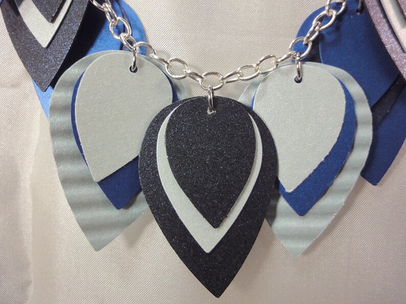 May include: A silver chain necklace with layered teardrop-shaped pendants. The pendants are made of blue, white, and black felt with a sparkly finish.