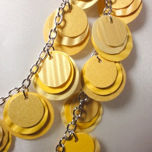 May include: A silver chain necklace with multiple layers of yellow paper circles. The circles are layered on top of each other to create a cascading effect.