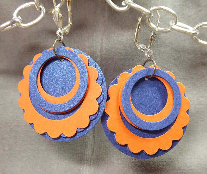 May include: A pair of clip-on earrings with three layers of circles. The outer layer is orange with scalloped edges, the middle layer is blue, and the inner layer is a smaller blue circle.