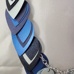 May include: A layered necklace made of blue, black, and silver teardrop-shaped pieces. The necklace is connected by a silver chain.