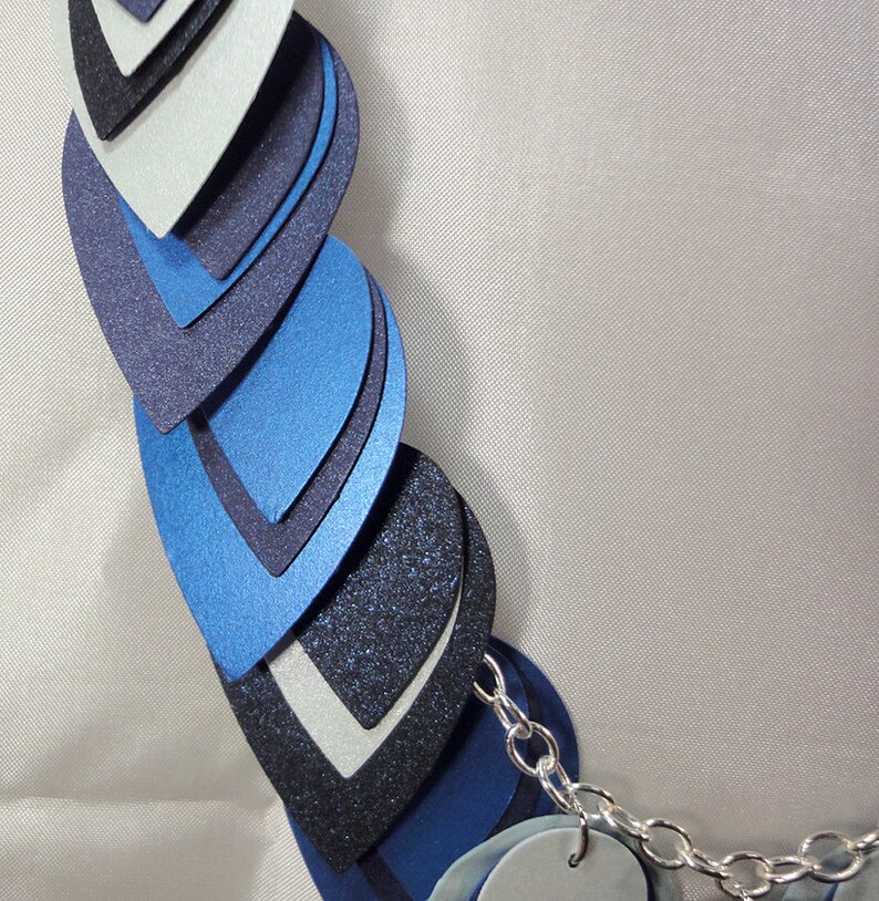 May include: A layered necklace made of blue, black, and silver teardrop-shaped pieces. The necklace is connected by a silver chain.