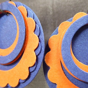 May include: A pair of earrings made of layered felt circles. The circles are orange and blue, with the blue circles having a scalloped edge.