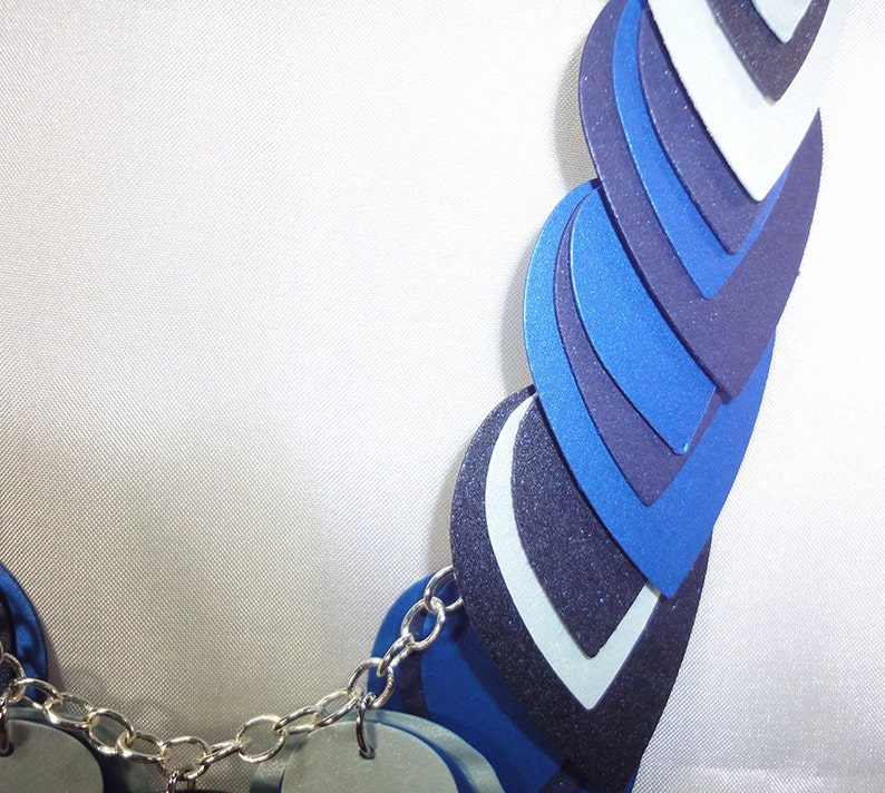 May include: A blue and white layered necklace with a silver chain. The necklace is made of multiple layers of blue and white fabric, each layer is a different shade of blue. The necklace is a statement piece and would be perfect for a special occasion.