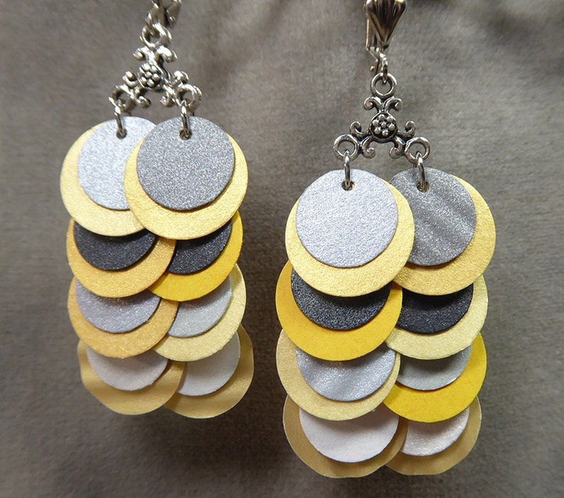 K&ouml;nnte beinhalten: Ein Paar Ohrringe mit mehreren Schichten runder Scheiben in Silber, Gold und Schwarz. Die Scheiben sind in einem abgestuften Muster &uuml;bereinander gelegt, wobei die gr&ouml;&szlig;te Scheibe oben und die kleinste unten ist. Die Ohrringe sind an einem silbernen Haken befestigt.