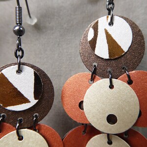 May include: A pair of dangle earrings with multiple round discs. The discs are layered and feature a brown, white, and orange color scheme. The top disc has a geometric pattern in brown and white.