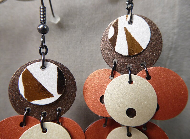 May include: A pair of dangle earrings with multiple round discs. The discs are layered and feature a brown, white, and orange color scheme. The top disc has a geometric pattern in brown and white.