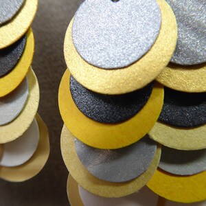 K&ouml;nnte beinhalten: Ein Paar Ohrringe mit mehreren Schichten runder Scheiben in Gold, Silber und Schwarz. Die Scheiben sind in abgestufter Gr&ouml;&szlig;e &uuml;bereinander gelegt, wobei die gr&ouml;&szlig;te Scheibe oben und die kleinste Scheibe unten ist.