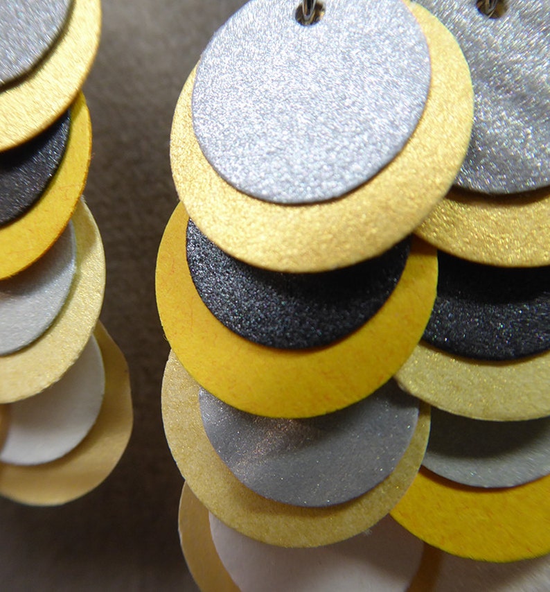 K&ouml;nnte beinhalten: Ein Paar Ohrringe mit mehreren Schichten runder Scheiben in Gold, Silber und Schwarz. Die Scheiben sind in abgestufter Gr&ouml;&szlig;e &uuml;bereinander gelegt, wobei die gr&ouml;&szlig;te Scheibe oben und die kleinste Scheibe unten ist.