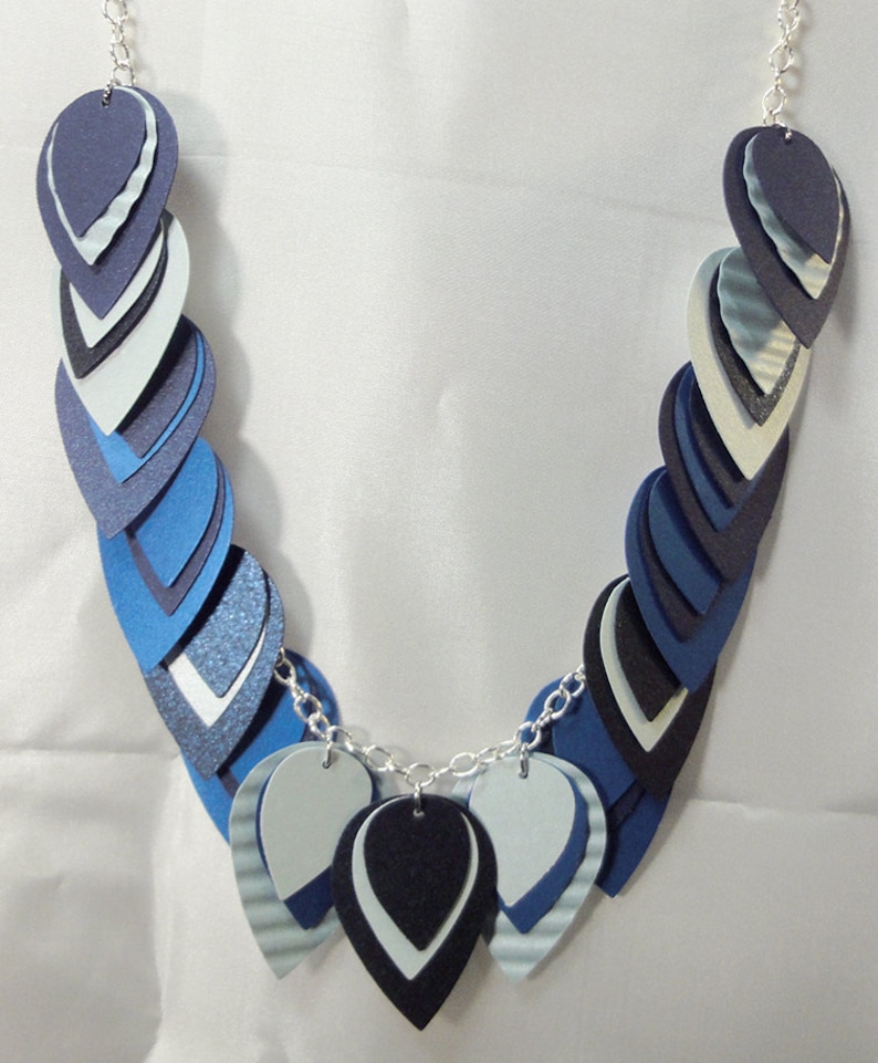 May include: A necklace made of layered blue, white, and black paper teardrop shapes. The necklace is strung on a silver chain.